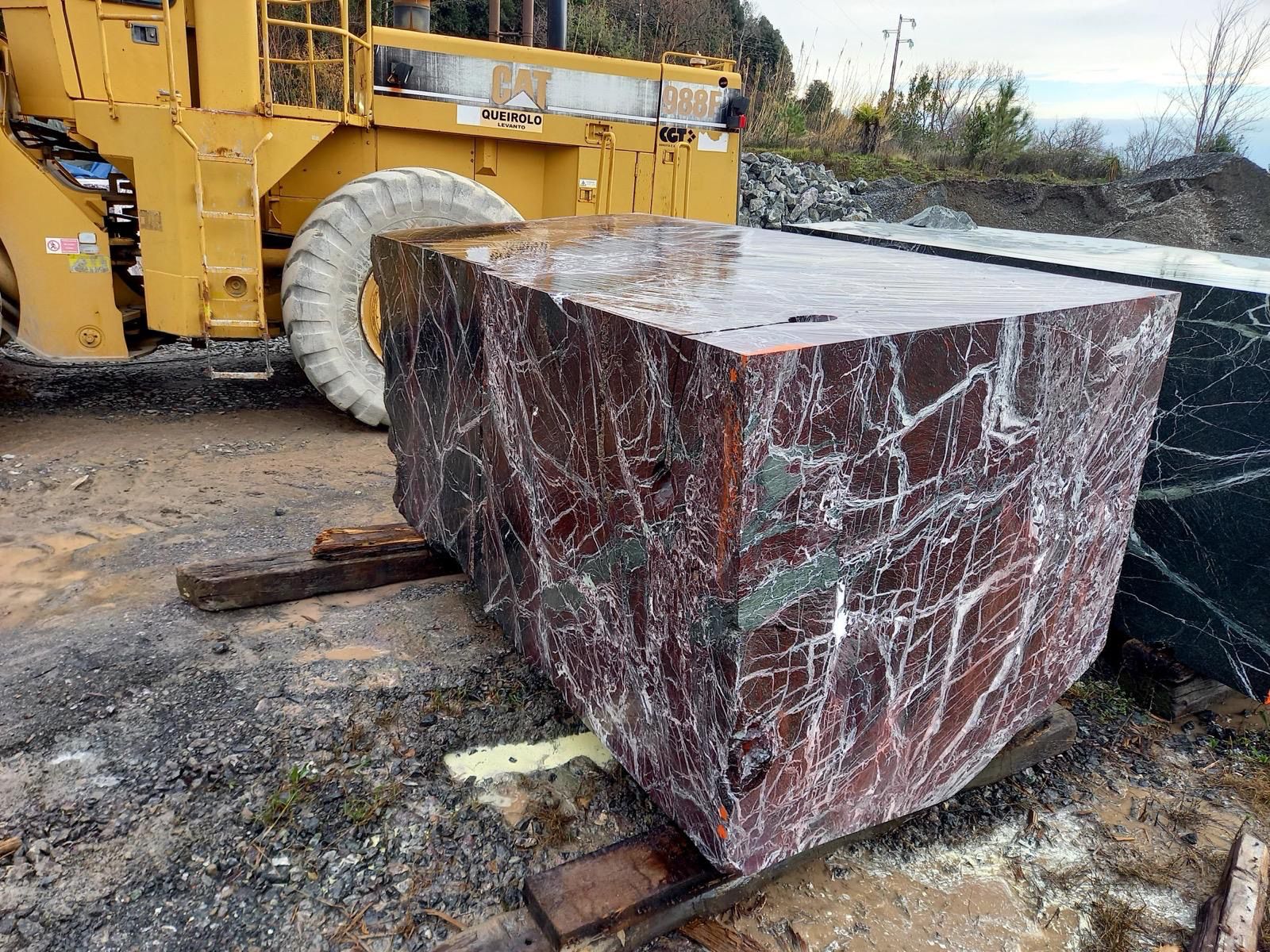 Large raw Rosso Levanto marble block cut and squared in the quarry yard, with heavy equipment in the background