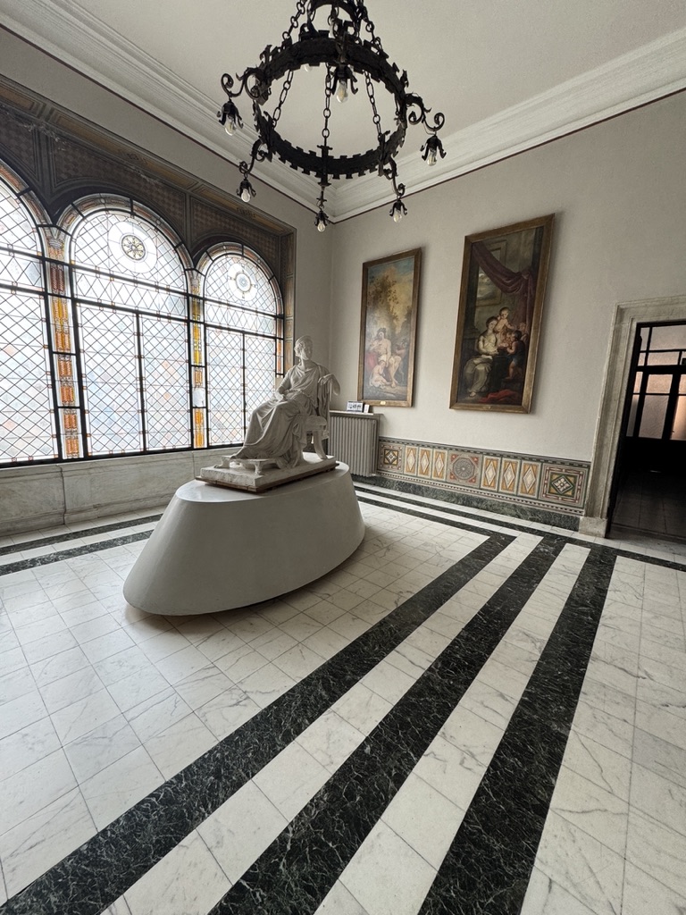 Grand marble hall with a seated classical sculpture on a curved plinth, stained-glass arched windows, and a black wrought-iron chandelier over a black-and-white striped marble floor