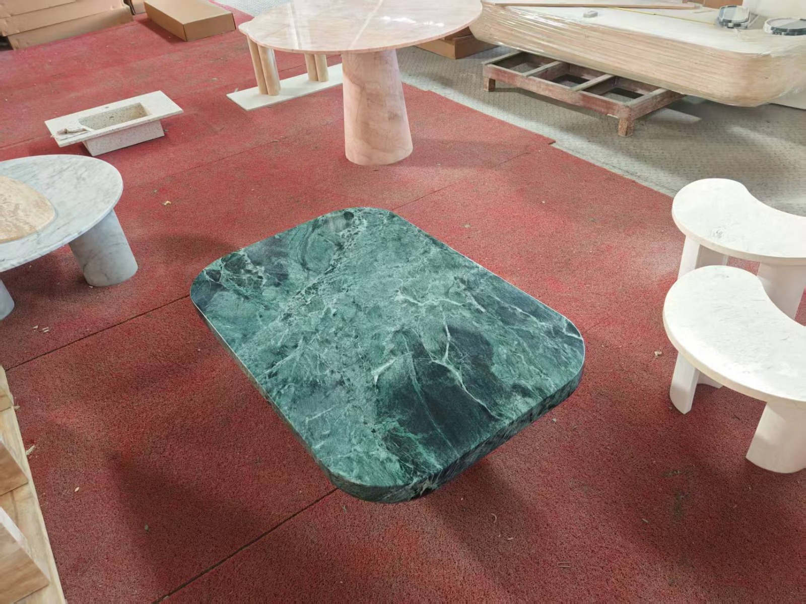 Low rounded-rectangle coffee table in deep green marble with white veining, on a red workshop floor surrounded by other stone pieces