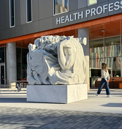 Monumental sleeping marble giant installed on a plaza in front of a Health Professions building, passer-by giving sense of scale