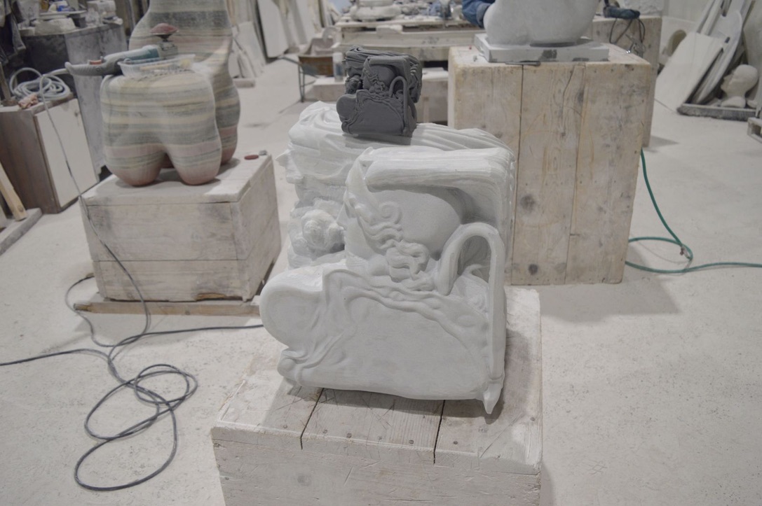 White marble sculpture in progress on a wooden plinth in the workshop, with a small grey maquette resting on top