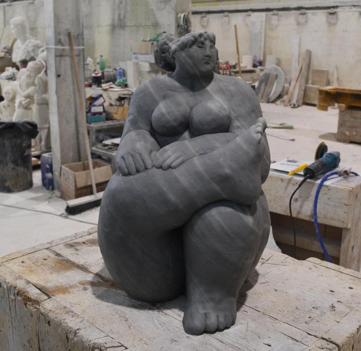 Grey marble sculpture of a seated voluptuous female figure holding her foot, front view in the workshop