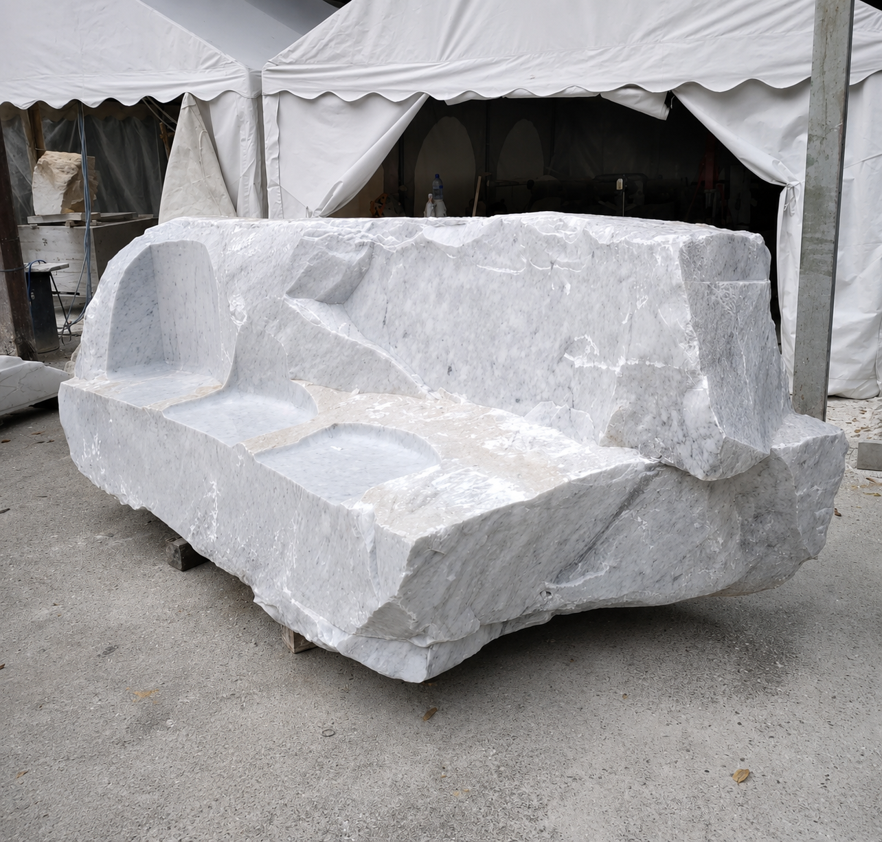 Monumental Carrara marble block carved into a rough-hewn sofa with smooth seat hollows, displayed under a white tent