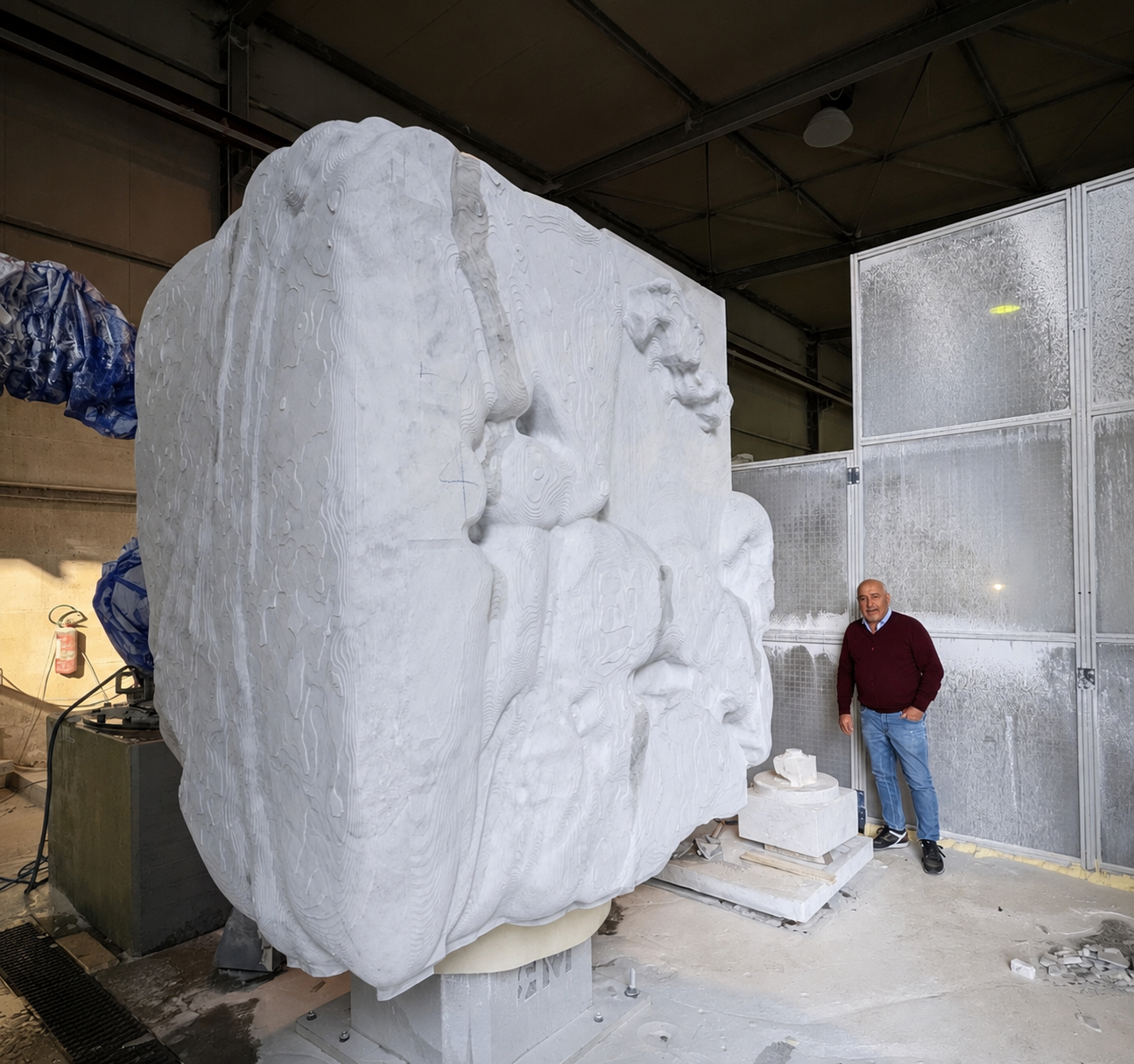 Monumental white marble cube carved with emerging faces and bodies, shown beside a man for scale in the workshop