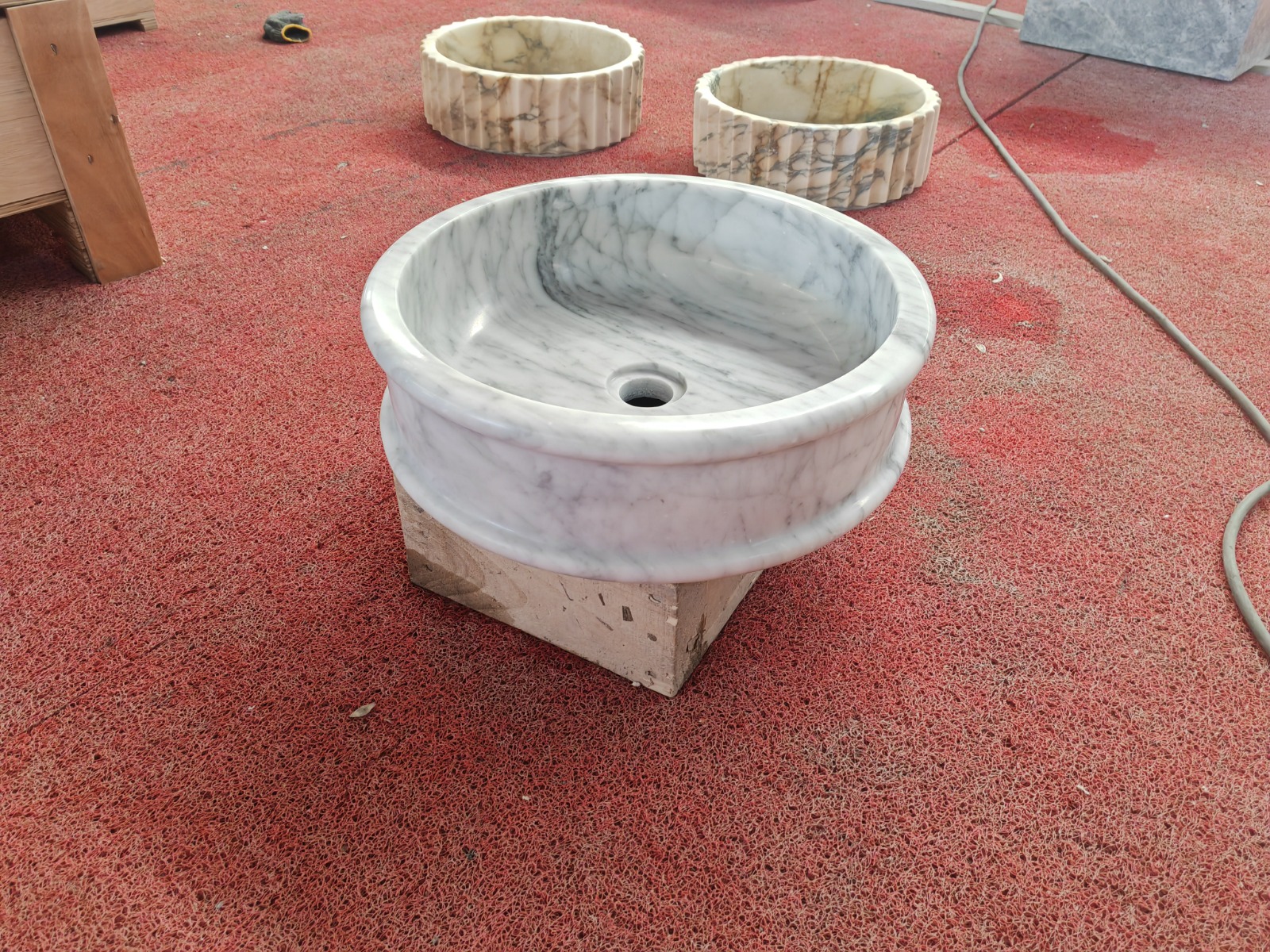 Round Carrara marble vessel sink resting on a travertine base, with two fluted Calacatta sinks behind on a red workshop floor