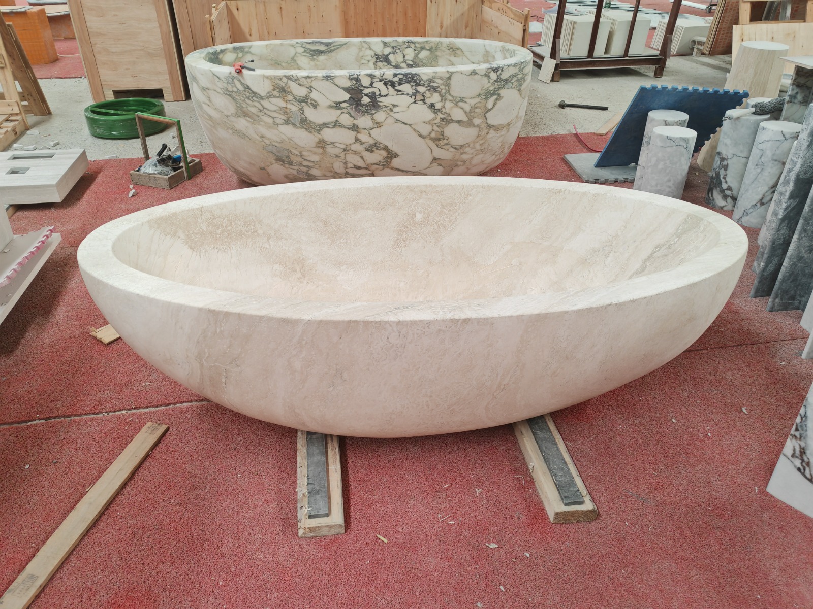 Two freestanding oval marble bathtubs in the workshop — one in cream travertine and one in Calacatta Viola