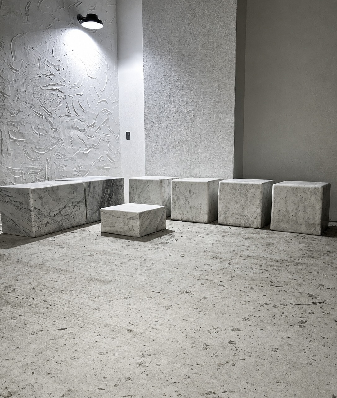 Row of seven solid Carrara marble cube seats arranged in a gallery space with a textured plaster wall