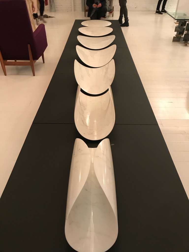 Row of seven Carrara marble pod sculptures aligned on a long dark plinth in a gallery