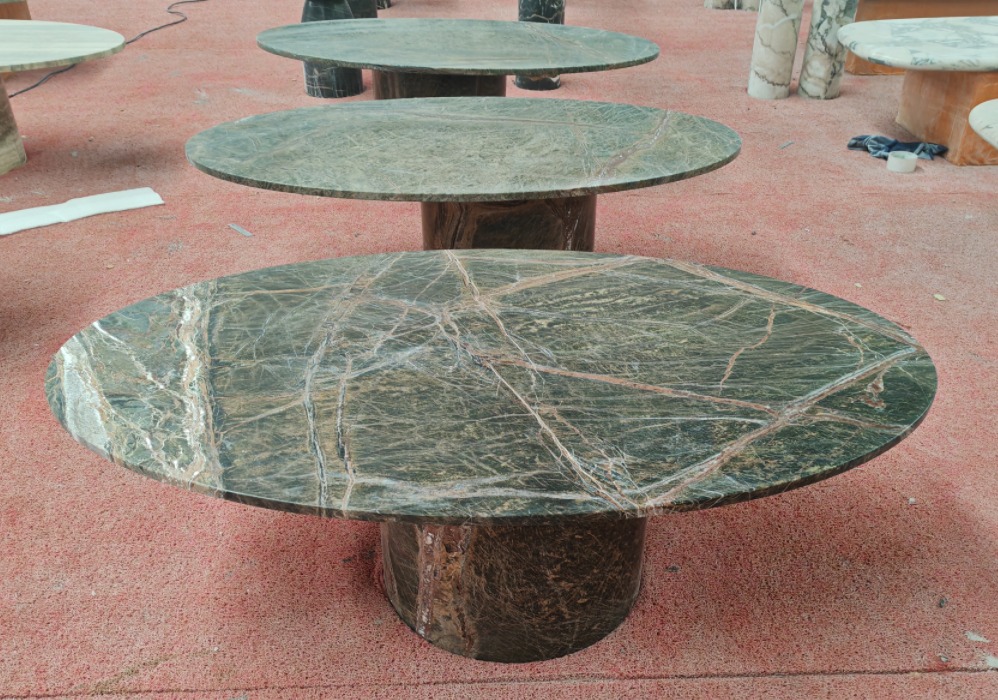 Three large oval Verde marble coffee tables with deep green, white, and rust veining on cylindrical pedestals, lined up on a red workshop floor