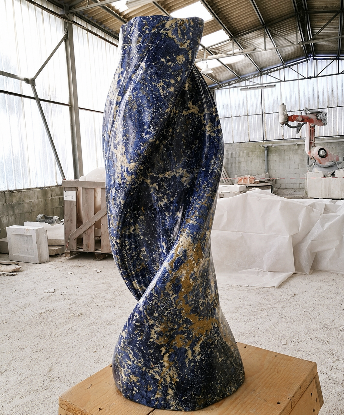 Tall twisted sculpture in deep blue sodalite with golden veining, standing on a wooden pallet in the workshop