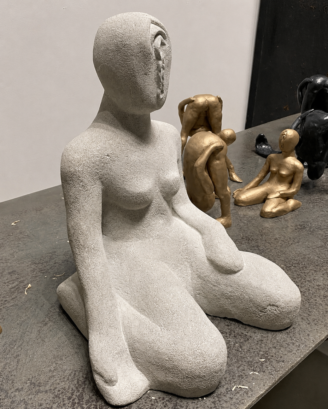 Textured seated white marble figure with bronze figures behind it on a workshop table