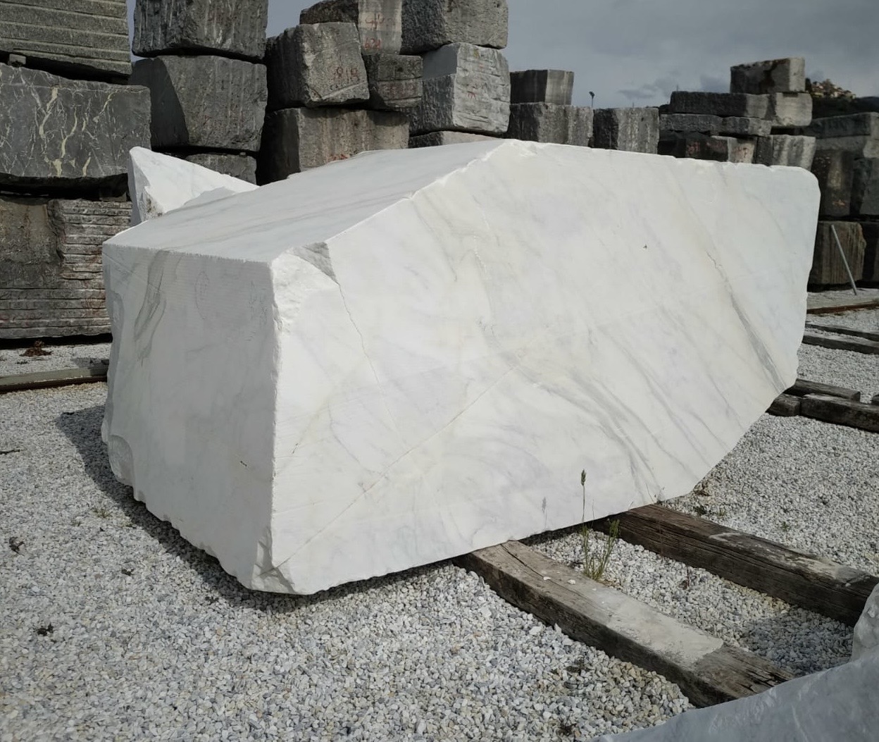 Freshly quarried raw block of Carrara White marble on wooden beams, with stacked grey blocks in the background